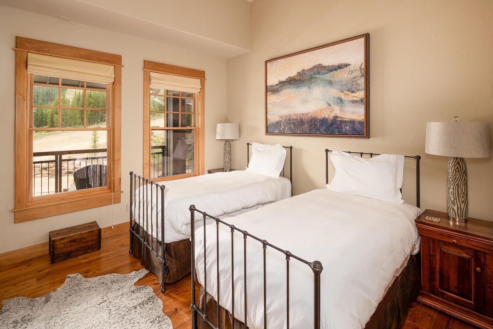 A bedroom with two beds, wooden floors, a large window, and a painting on the wall.