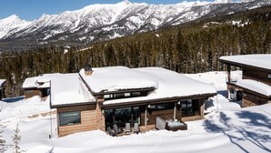Un chalet moderne doté d’une grande terrasse et d’un bain à remous, entouré de montagnes enneigées et d’une forêt dense.