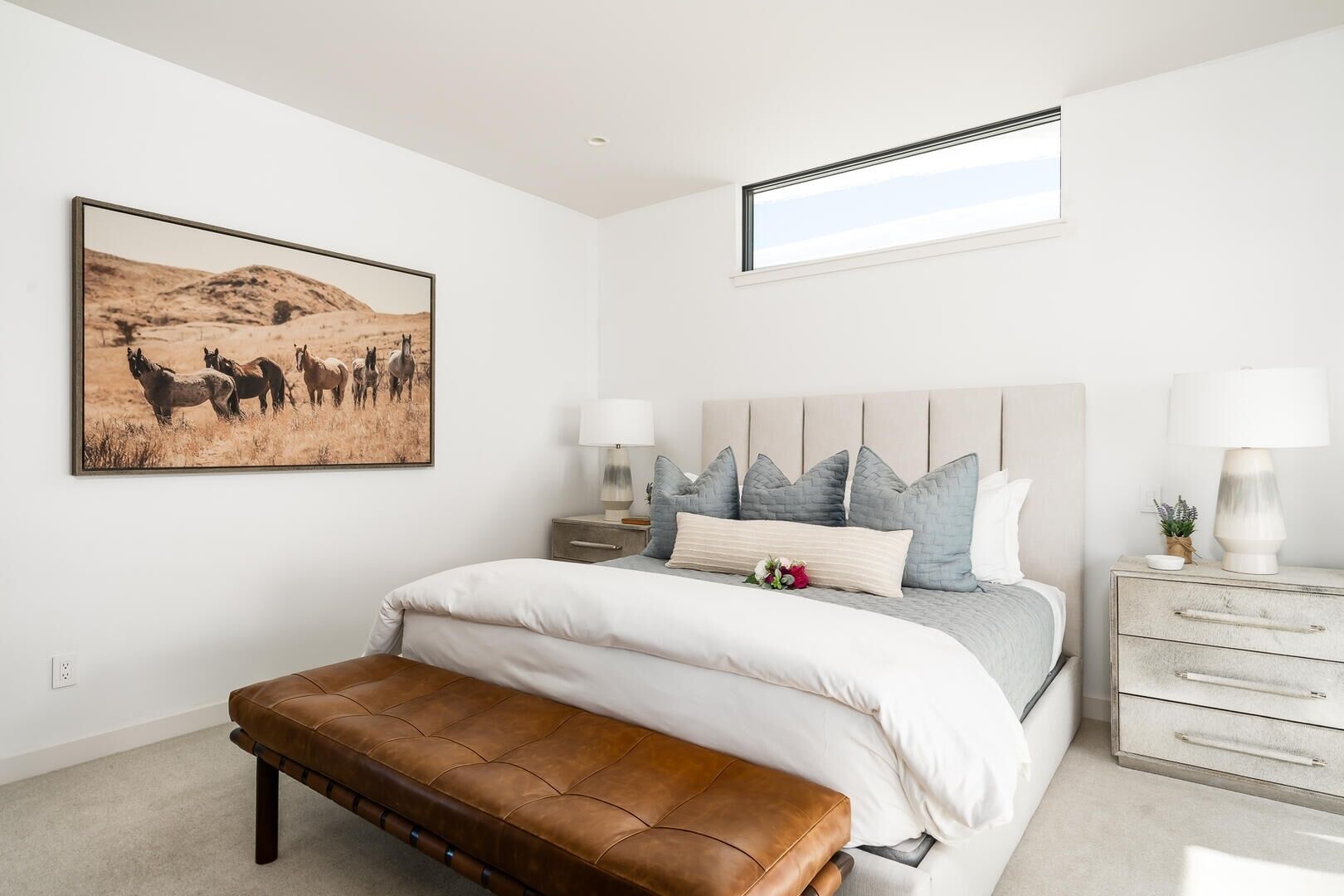 Een slaapkamer met een groot bed, een houten bankje, een nachtkastje, een dressoir, een raam en een schilderij van paarden.
