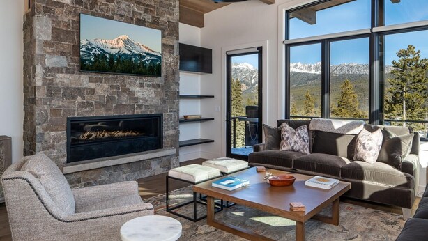 A modern living room with a stone fireplace, large windows, and a cozy seating area. - 9 Deep Forest Drive 6 Bedroom Home by Moonlight Basin Lodging (Big Sky)