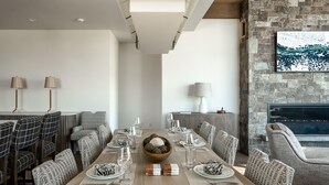 A modern dining room with a long table set for six, surrounded by patterned chairs. A fireplace and a stone wall are visible in the background. - 9 Deep Forest Drive 6 Bedroom Home by Moonlight Basin Lodging (Big Sky)