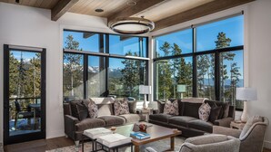 A spacious living room with large windows, a wooden ceiling, and a cozy seating area with a coffee table. - 9 Deep Forest Drive 6 Bedroom Home by Moonlight Basin Lodging (Big Sky)