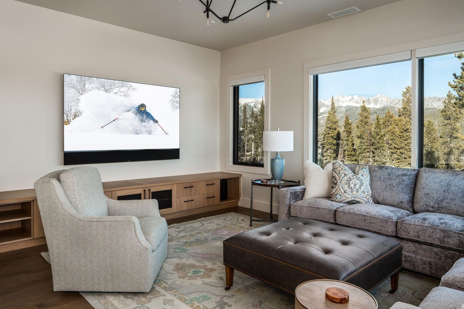 A modern living room with a large flat-screen TV, a grey armchair, a brown leather ottoman, a grey sofa with patterned cushions, and a wooden entertainment unit.