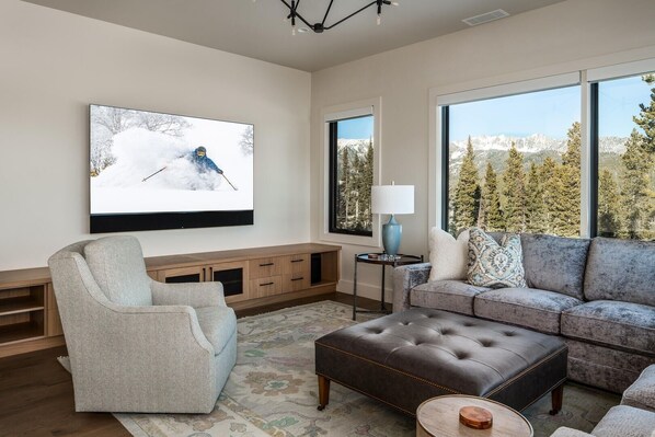 A modern living room with a large flat-screen TV, a grey armchair, a brown leather ottoman, a grey sofa with patterned cushions, and a wooden entertainment unit.