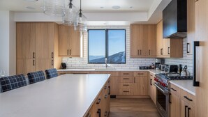 A modern kitchen with wooden cabinets, a large island, and a view of a snowy landscape. - 9 Deep Forest Drive 6 Bedroom Home by Moonlight Basin Lodging (Big Sky)