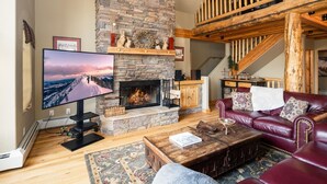 A cozy living room with a stone fireplace, a television, a leather sofa, and a staircase. - 6 Mountain Home Road 4 Bedroom Home by Moonlight Basin Lodging (Big Sky)