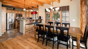 A spacious kitchen with wooden cabinets and a central island, a dining area with a large wooden table and chairs, and a chandelier. - 6 Mountain Home Road 4 Bedroom Home by Moonlight Basin Lodging (Big Sky)