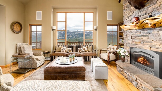 A cozy living room with a stone fireplace, a large window overlooking mountains, and a mounted animal head on the wall. - 3 Bandit Way 4 Bedroom Home by Moonlight Basin Lodging (Big Sky)
