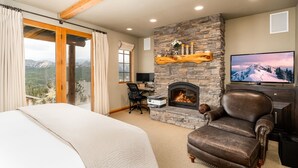 A bedroom with a stone fireplace, a large bed, a leather armchair, and a desk with a computer. - 3 Bandit Way 4 Bedroom Home by Moonlight Basin Lodging (Big Sky)
