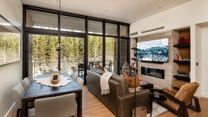 A modern living room with a dining area, large windows, a fireplace, and a TV. - 215 Lakelodge 2 Bedroom Condo by Moonlight Basin Lodging (Big Sky)