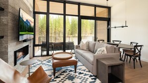 A modern living room with a fireplace, large windows, a sofa, a dining table, and a TV mounted on the wall. - 221 Lakelodge 2 Bedroom Condo by Moonlight Basin Lodging (Big Sky)