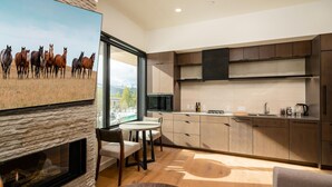A modern kitchen with a stone fireplace, wooden cabinets, and a large window overlooking a field. - 223 Lakelodge 1 Bedroom Condo by Moonlight Basin Lodging (Big Sky)