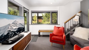 A modern living room with a large flat-screen TV, a red chair, a grey sofa, and a staircase with a white lamp. - 27 Lake Cabin 3 Bedroom Home by Moonlight Basin Lodging (Big Sky)