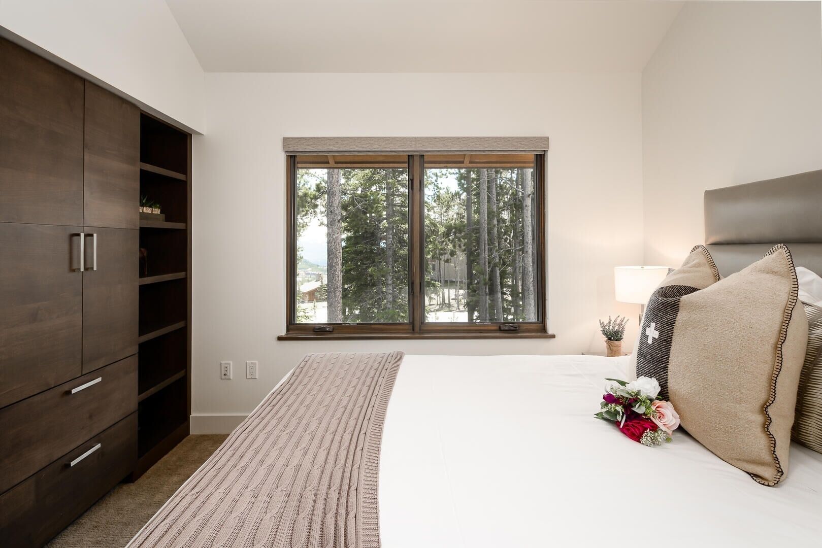 A bedroom with a large window, a bed with pillows, a bedside table with a lamp, and a wardrobe.