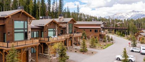 A row of log cabin-style buildings with balconies, surrounded by trees and a mountain backdrop.