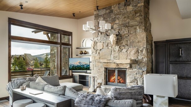 A modern living room with a stone fireplace, a large window with a view of the mountains, and a cozy seating area. - 4b Lodgeside 3 Bedroom Townhouse by Moonlight Basin Lodging (Big Sky)