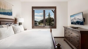 A hotel room with a large bed, a dresser, a television, and a window with a mountain view. - 4b Lodgeside 3 Bedroom Townhouse by Moonlight Basin Lodging (Big Sky)