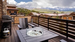 A wooden deck with a glass-top table and chairs, a propane grill, and a fire pit. - 4b Lodgeside 3 Bedroom Townhouse by Moonlight Basin Lodging (Big Sky)