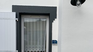 Une porte blanche ornée d’un rideau en dentelle, une boîte aux lettres noire et une plaque bleue portant le numéro 18.