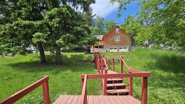 Eine Holzterrasse führt zu einem Haus mit steinernen Fundamenten, umgeben von üppiger Vegetation und Bäumen.