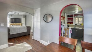 Ein Wohnzimmer mit einer Couch, einem Fernseher und einem Esstisch mit Stühlen. Durch einen Bogen ist eine Küche erkennbar.
