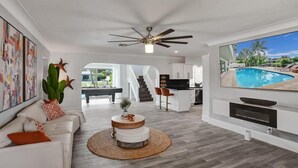 A modern living room with a white sofa, a round coffee table, and a ceiling fan. There is a kitchen area in the background with a staircase leading to an upper level.