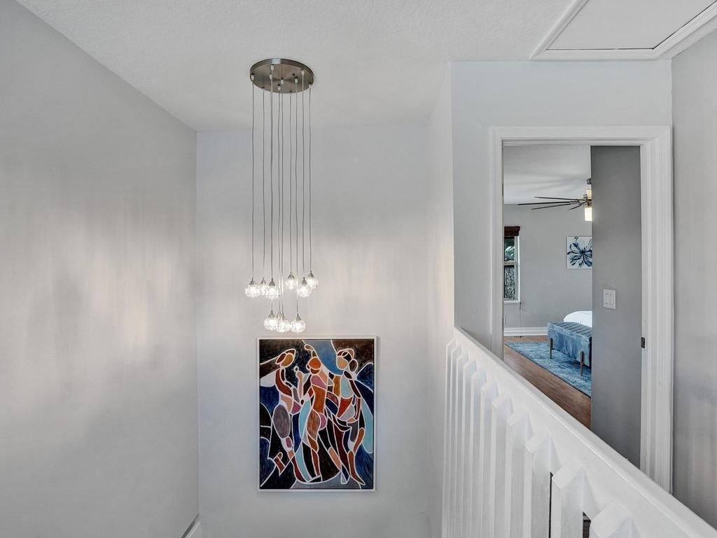 A hallway with a modern chandelier, a framed abstract painting, and a glimpse of a bedroom with a bed and a blue rug.