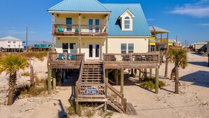 A two-story beach house with a blue roof, wooden deck, and a sign reading "Shore Beats Work". - Ocean view, steps from water, private bedroom balcony! - Shore Beats Work (Dauphin Island)