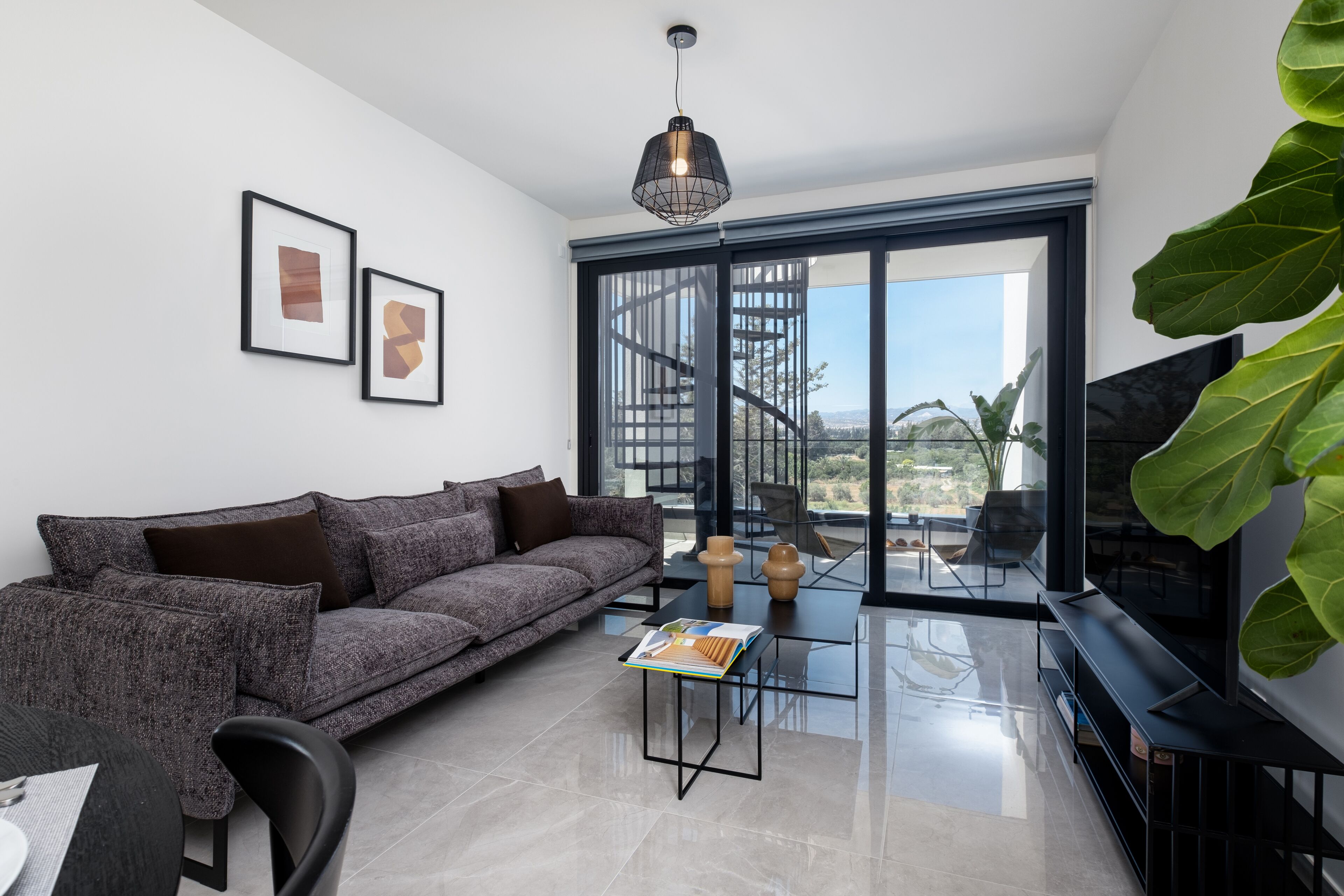 A modern living room with a grey sofa, a glass coffee table, a black TV stand, and a large window with a view of a spiral staircase and greenery.