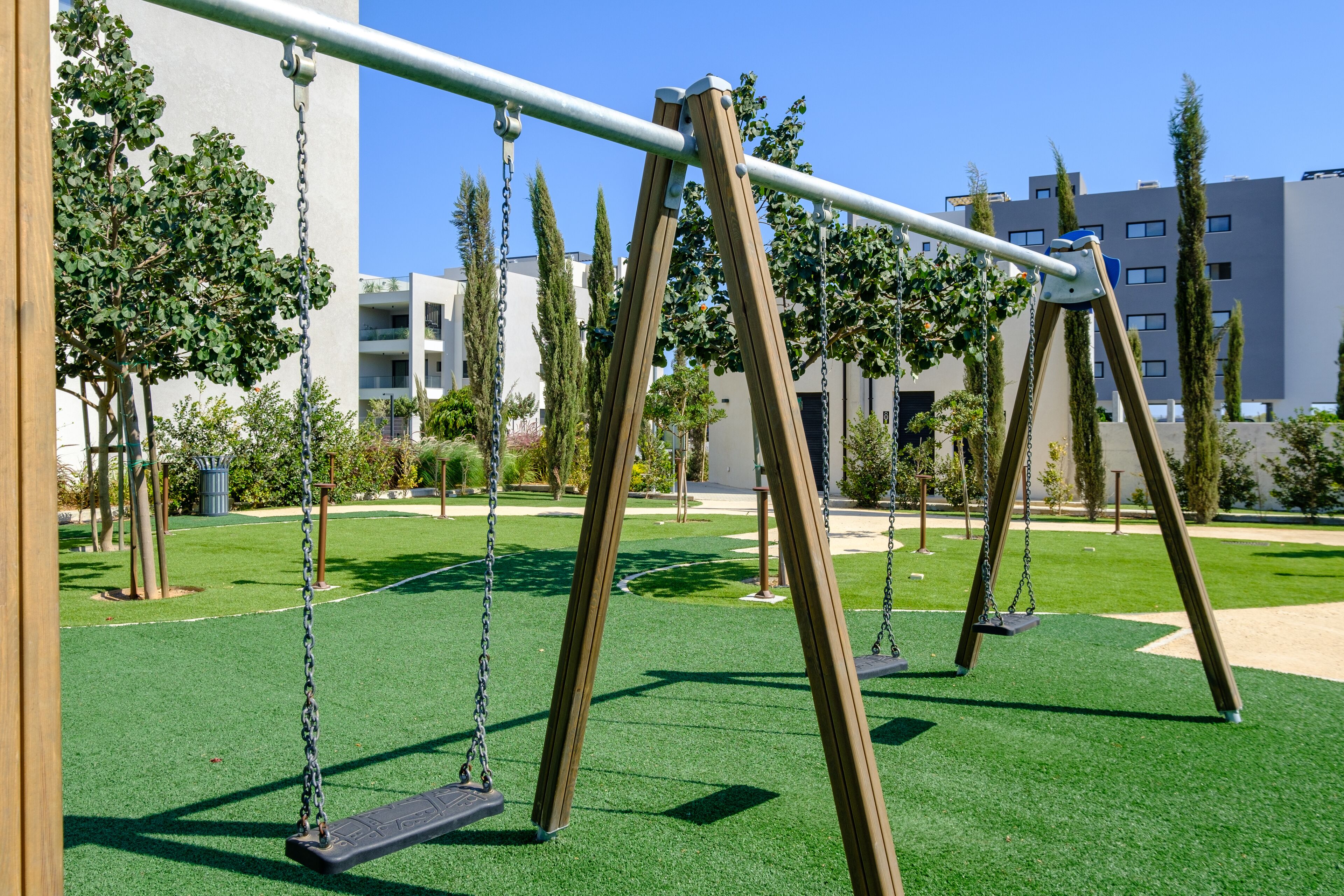 A playground with swings and artificial grass, surrounded by trees and buildings.
