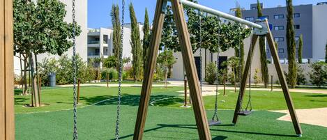 A playground with swings and artificial grass, surrounded by trees and buildings.
