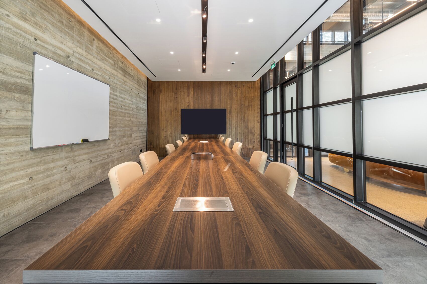 Une salle de conférence avec une longue table en bois, des chaises beiges, un tableau blanc et de grandes fenêtres.