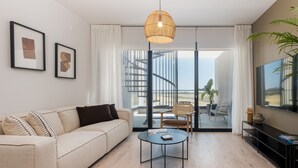 A modern living room with a sofa, coffee table, and a TV. There are framed pictures on the wall, a large window with curtains, and a staircase leading to a balcony. - Sunset Gardens (Tserkezoi)