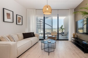 Una sala de estar moderna con sofá, mesa de centro y televisor. Hay cuadros enmarcados en la pared, un ventanal con cortinas y una escalera que lleva a un balcón.