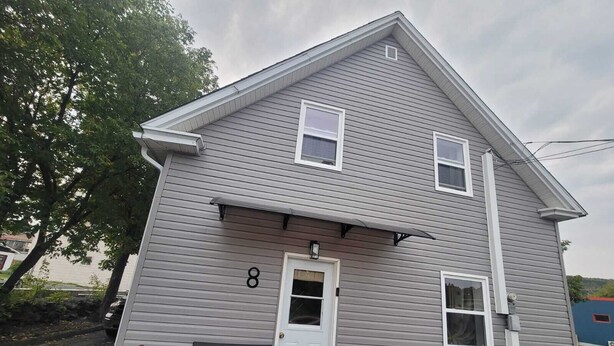 A two-story house with a grey siding exterior, a white door, and a small porch with a bench. - Bright 1 bedroom apartment close to highway, wifi, netflix, prime, free parking (Edmundston)