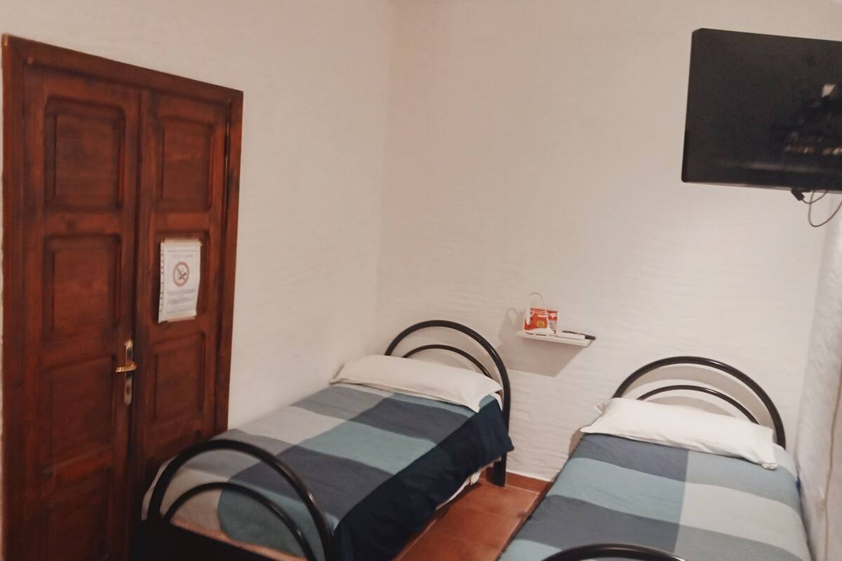 Una habitación pequeña con dos camas individuales, una puerta de madera, un televisor instalado en la pared y un estante con objetos.