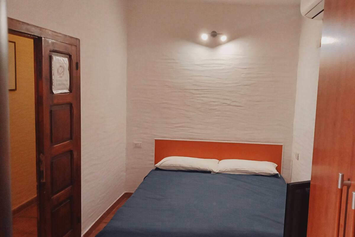 Una habitación pequeña con cama individual, puerta de madera, pared blanca y una lámpara en el techo.