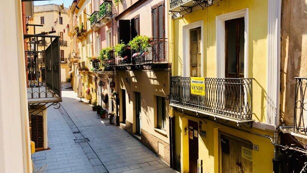 A narrow street with colorful buildings, balconies with plants, and a yellow building with a 'VENDE' sign.