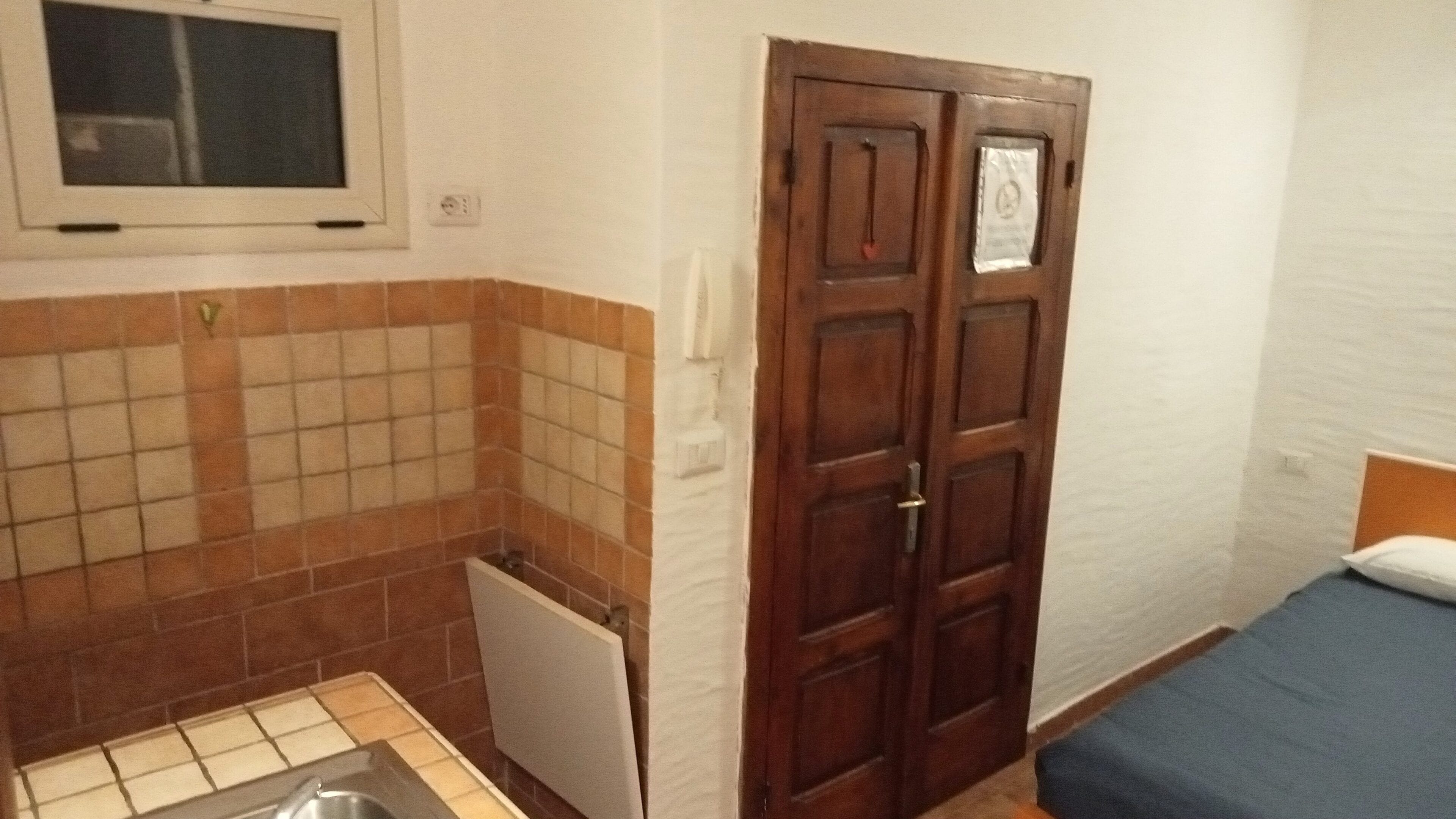 A small bathroom with a shower, a toilet, and a sink. There is a wooden door with a sign on it, and a bed with a blue blanket.