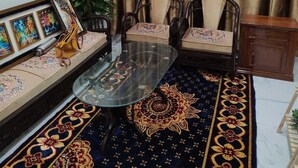 A living room with a glass coffee table, two armchairs, a sofa, a patterned rug, a small cabinet, and a potted plant.