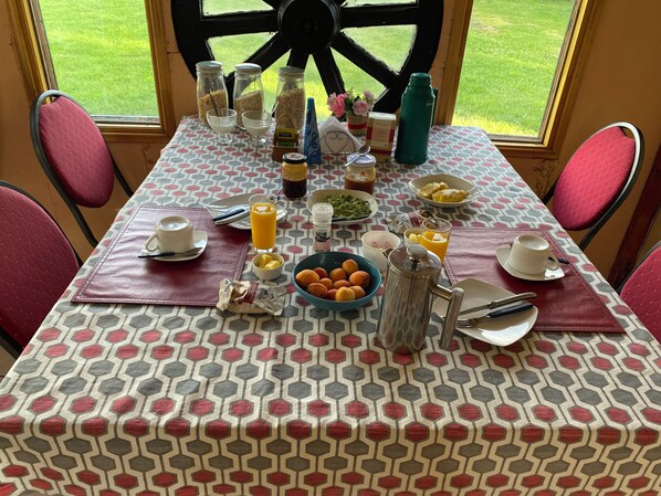 Mesa de desayuno preparada con diversos alimentos y bebidas, como cereal, jugo y pasteles, con vista a un jardín verde a través de un ventanal amplio.