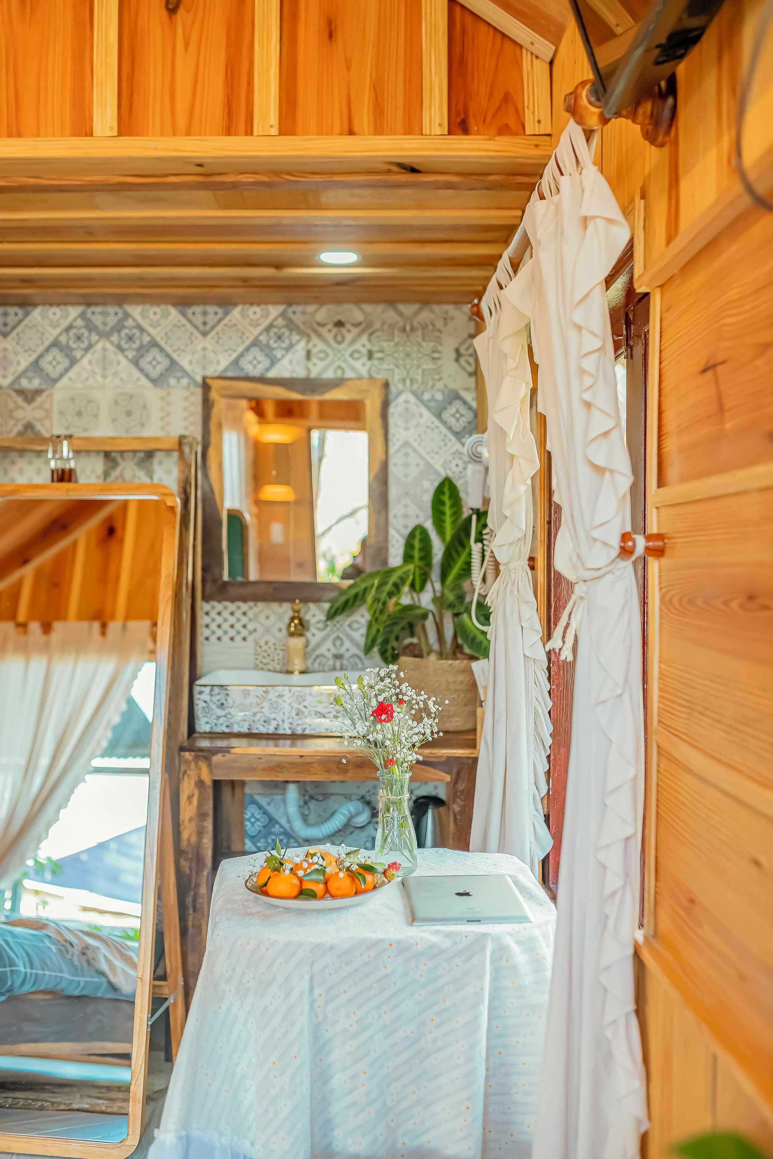 A compact room with a wooden interior, a small table with a laptop, a vase with flowers, and a window with curtains.