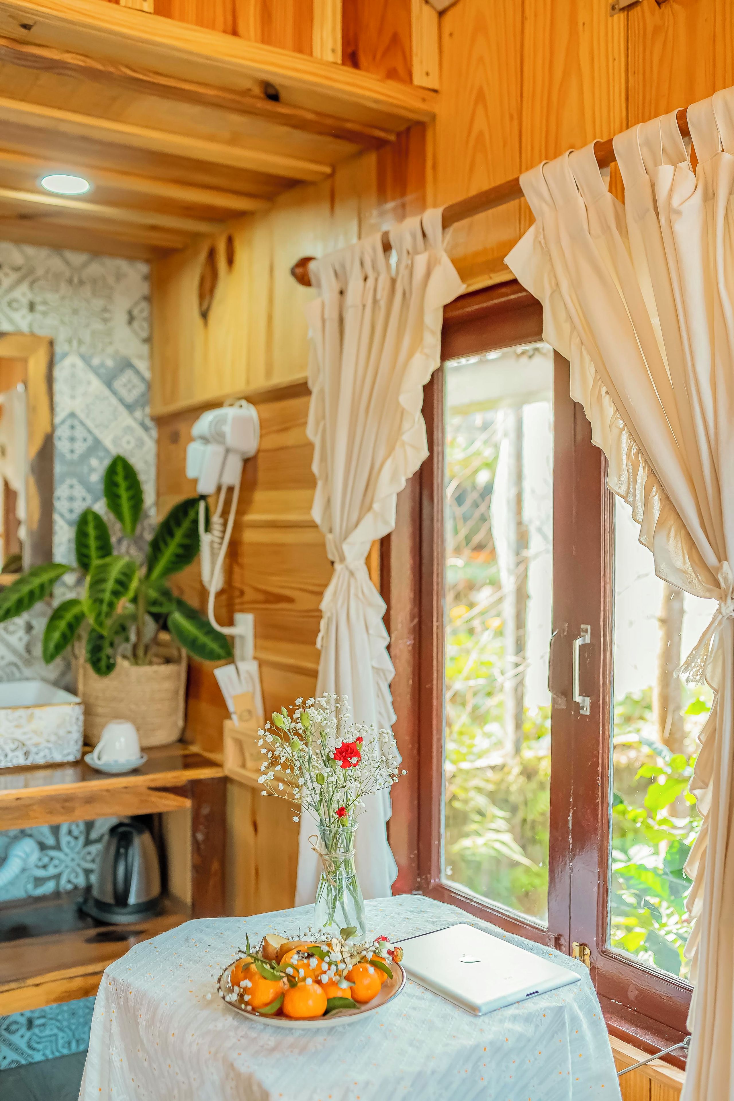 A wooden cabin interior with a table set for breakfast, a window with curtains, and a potted plant.