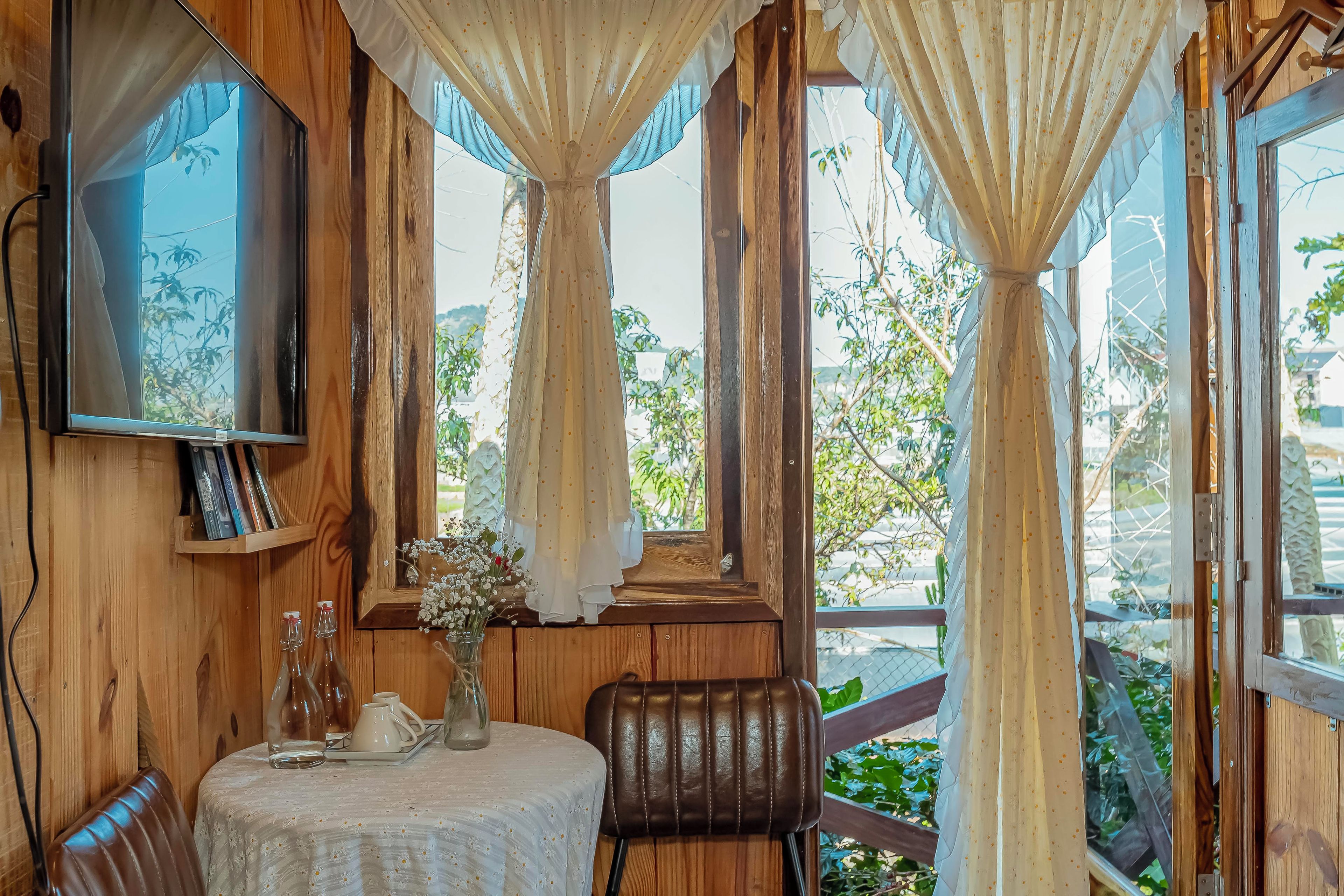A wooden room with a television, a table with a vase of flowers, and large windows with curtains.