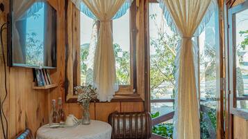A wooden room with a television, a table with a vase of flowers, and large windows with curtains.