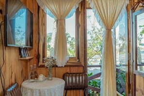 A wooden room with a television, a table with a vase of flowers, and large windows with curtains.