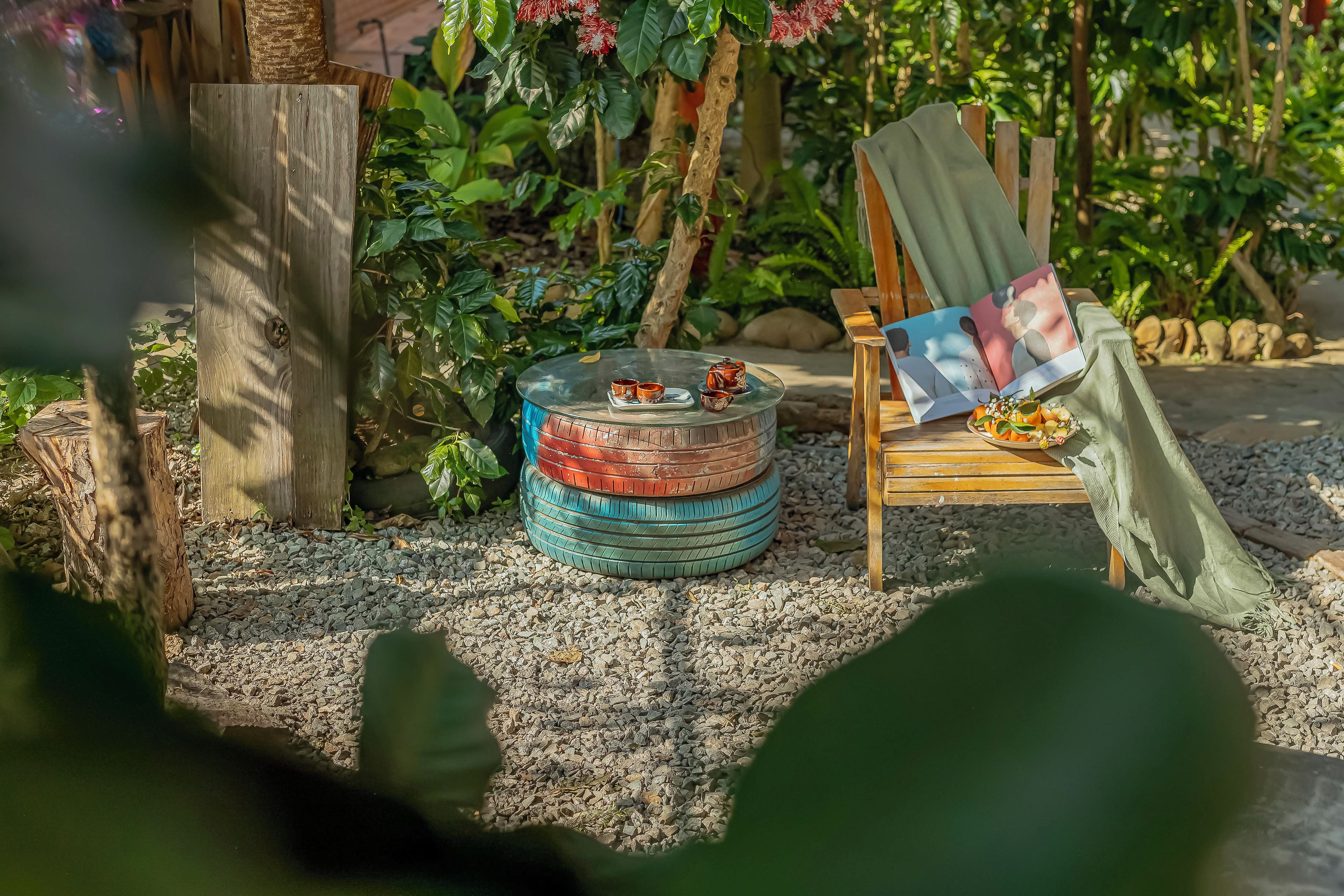 Um jardim com uma cadeira de madeira, mesa de vidro, um bule e chá, e um livro sobre a mesa.