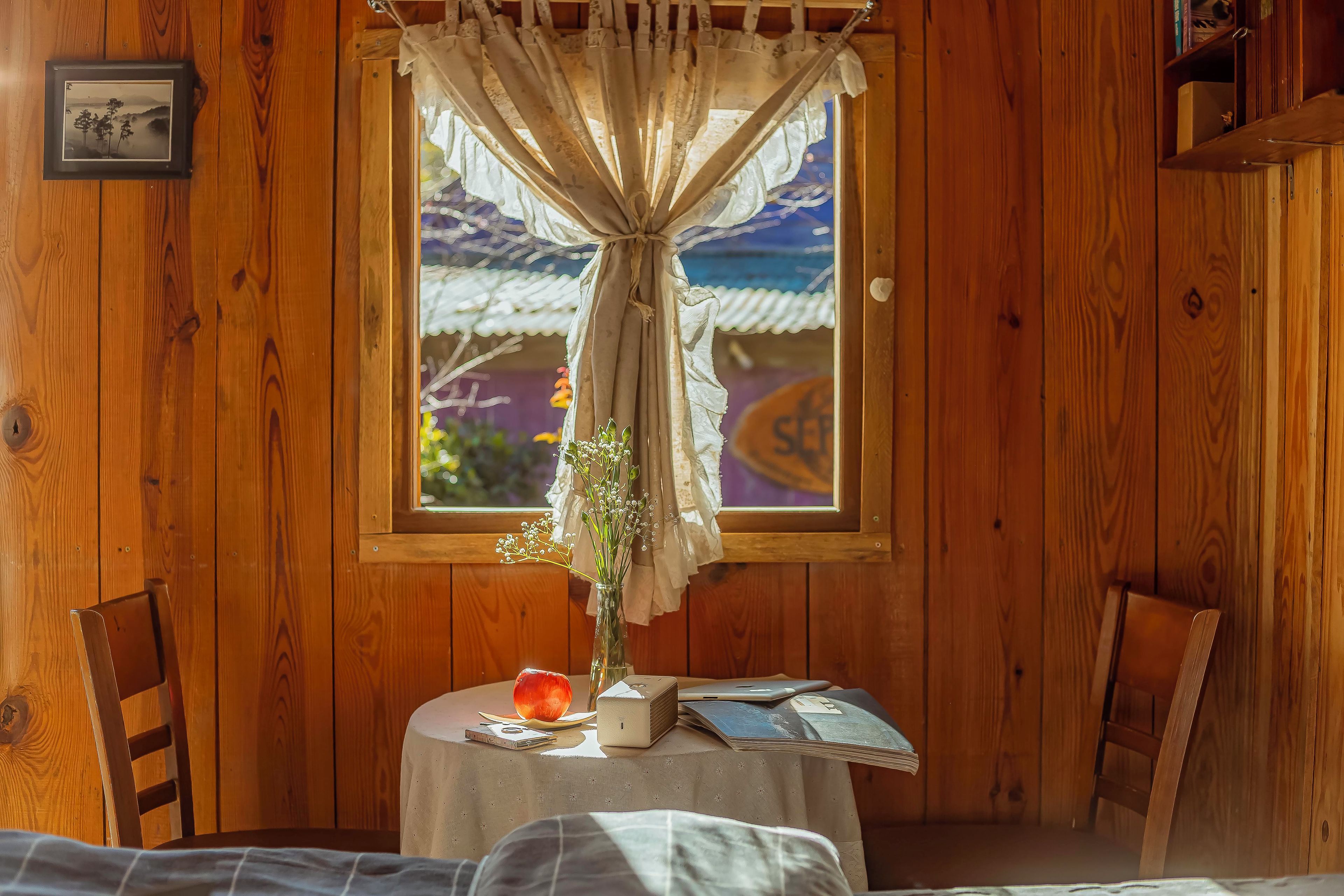 Um quarto de madeira com uma janela, uma mesa com um vaso de flores e uma cama.