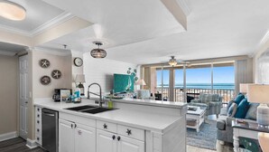 Une cuisine moderne dotée d’un îlot central, un coin repas offrant une vue sur la plage et un salon avec un balcon donnant sur l’océan.