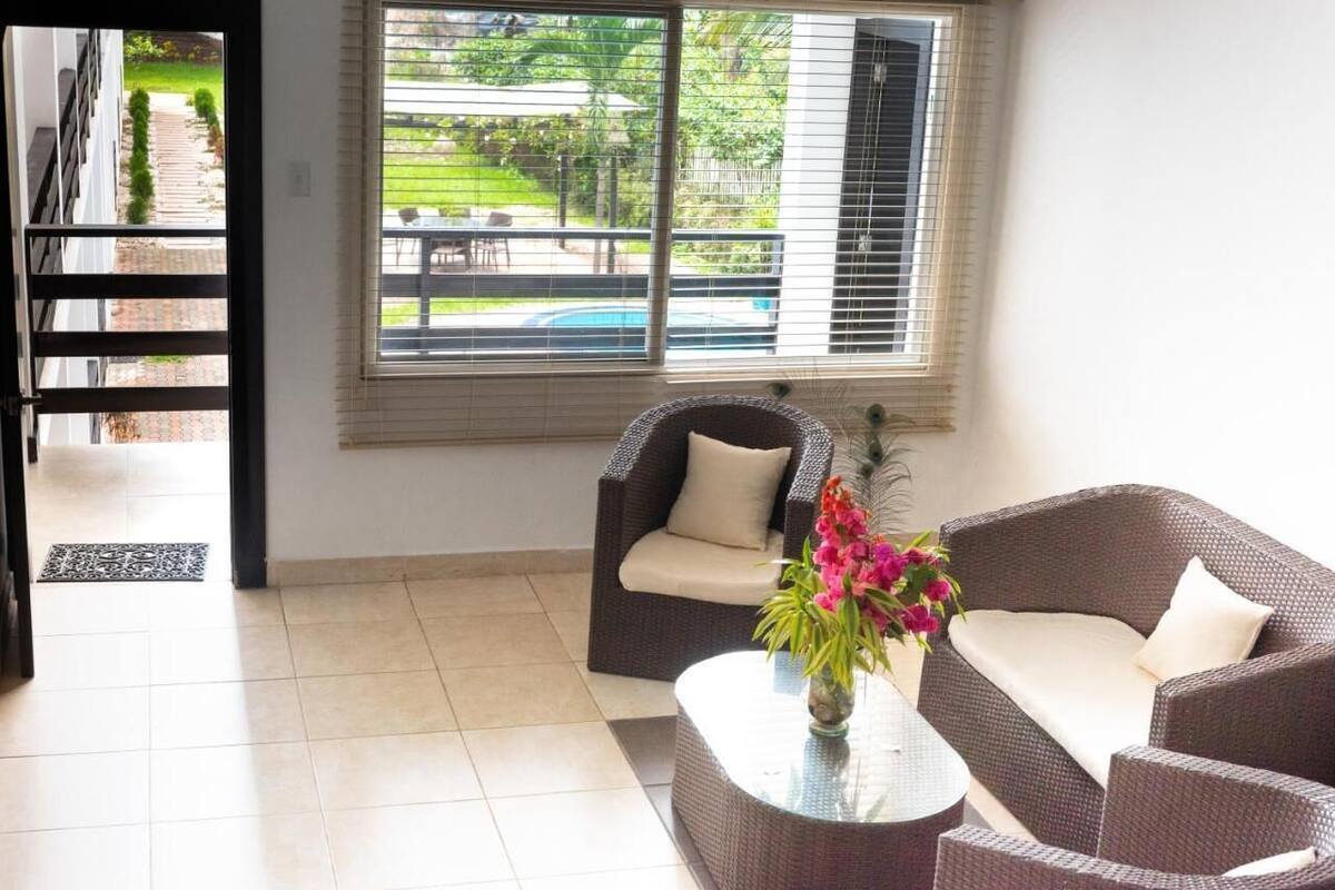 A room with a wicker sofa, a glass coffee table, and a view of a swimming pool and garden through large windows.